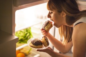 Close up image of a young woman with eating disorder, having a midnight snack - eating donuts, in front of the refrigerator.